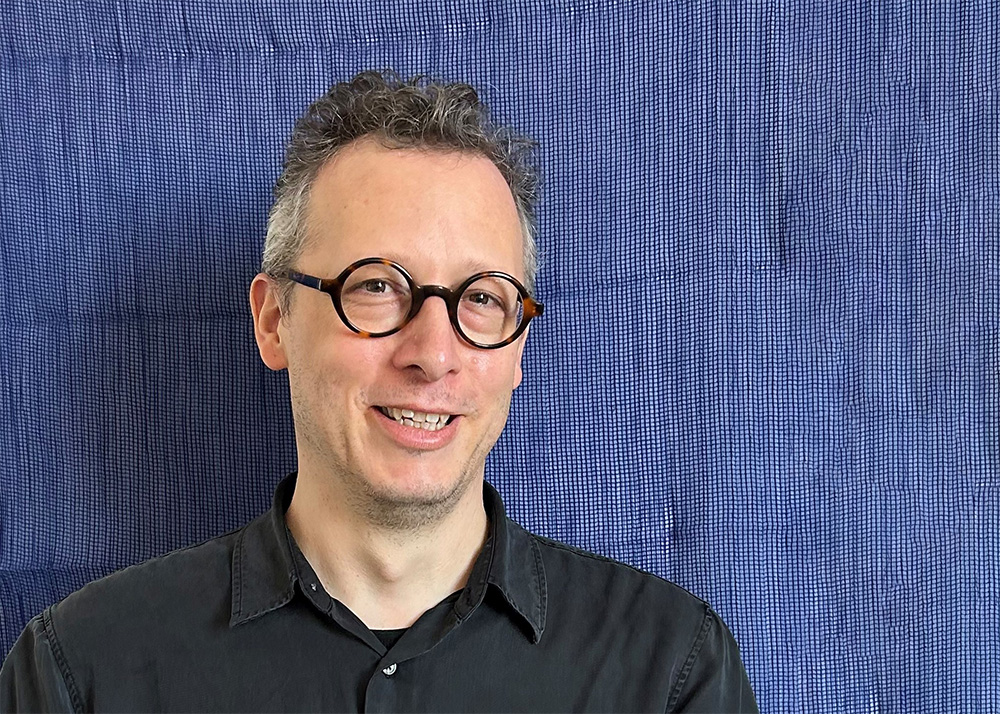 Porträttbild av Meriç Algün som står i grön skjorta framför en konstinstallation. Installationen är en vit vägg i ett ljust rum med citat skrivna i kursiv text.