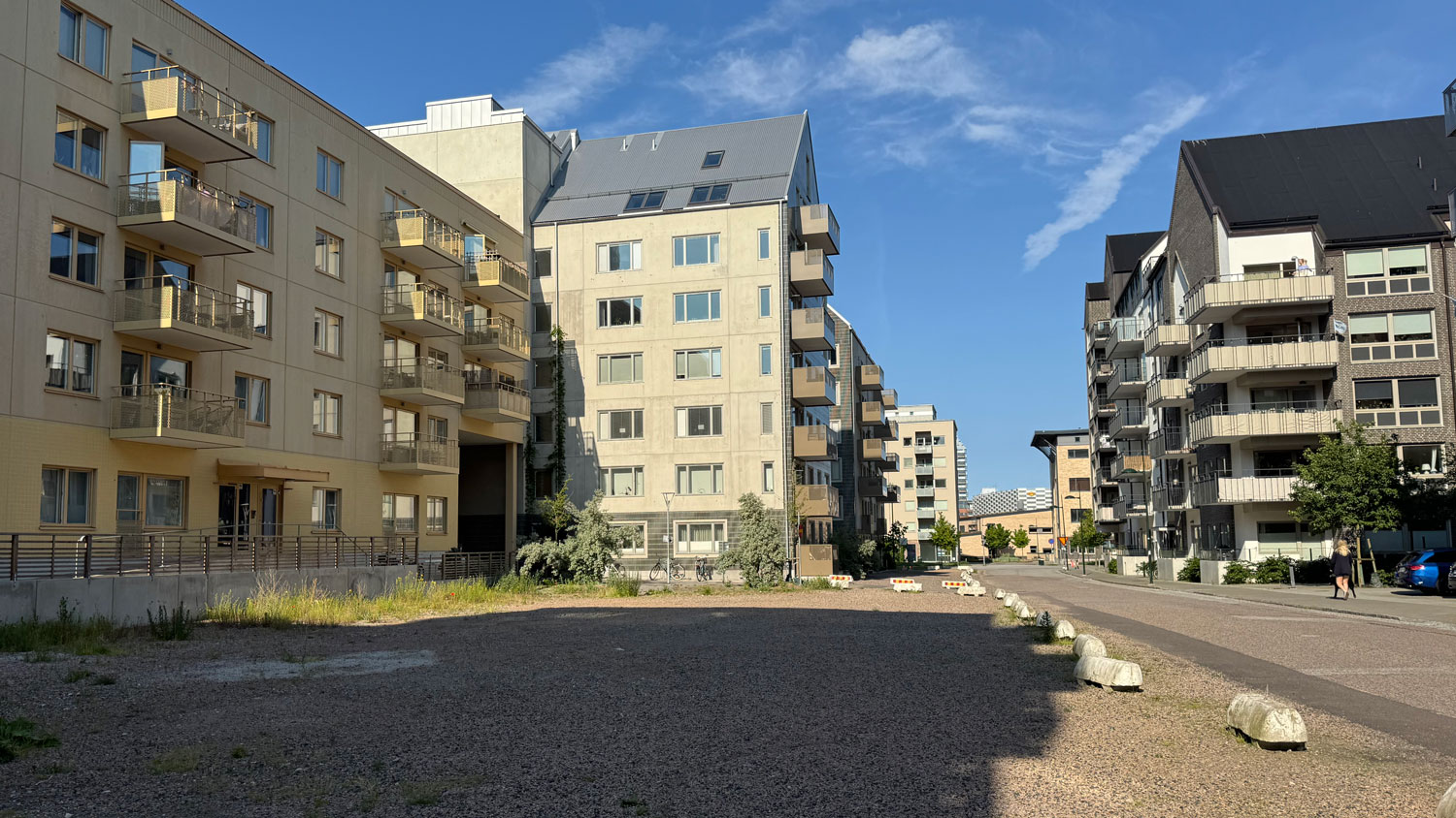 En grusad yta intill moderna bostadshus med guldgula och mörkgrå fasader, och låga betongklumpar som spärrar av mot gatan intill.