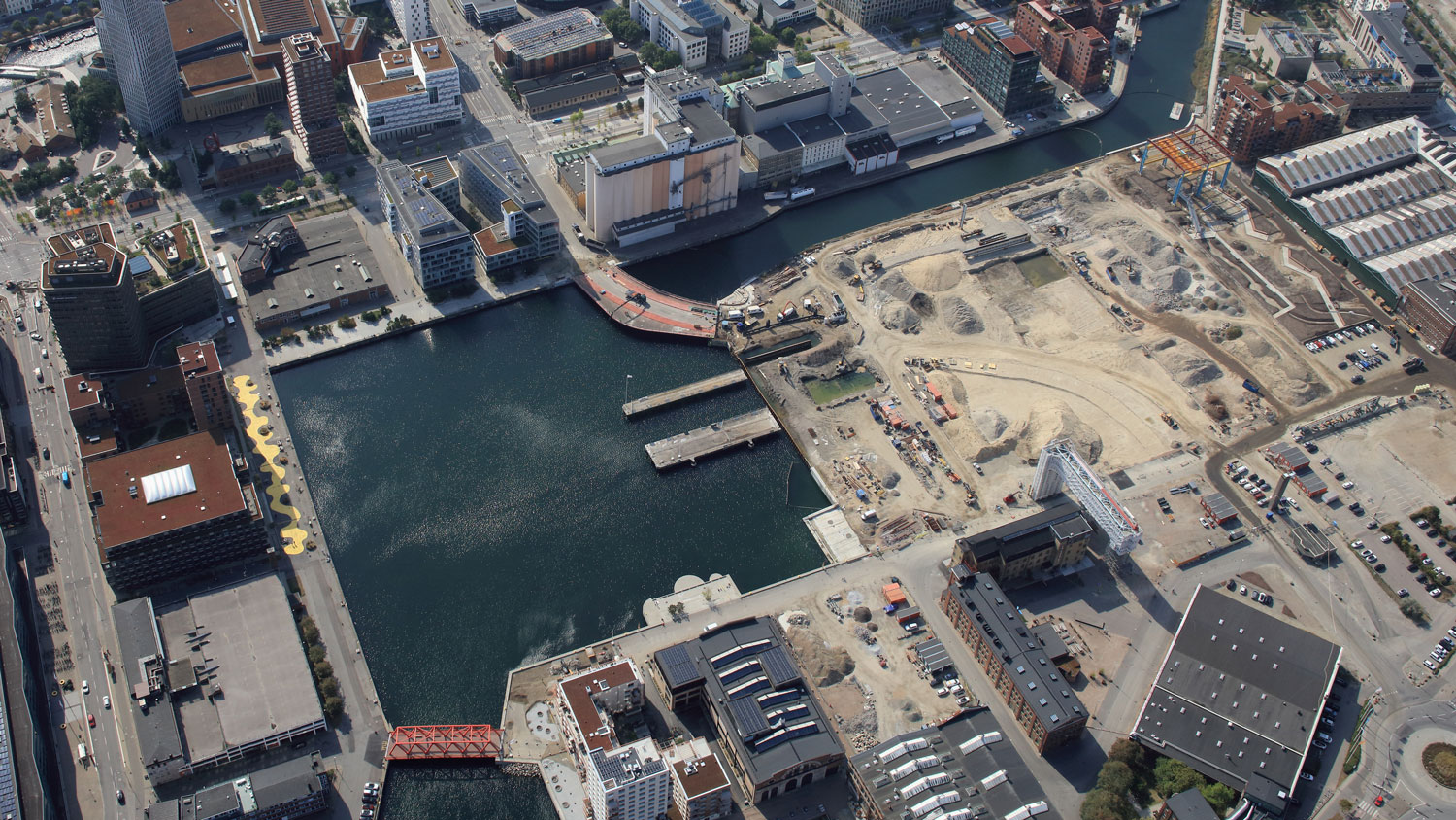 Flygvy över en kvadratisk hamnbassäng intill ett före detta varvsområde med varvskanal. Kajer, broar och byggnader runtom vattnet.