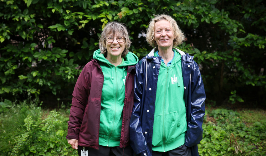 Åse och Anna står utomhus i grönskan i arbetskläder.