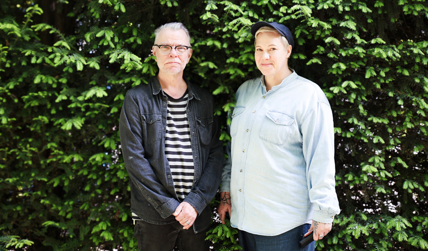 Peter och Anna står utomhus. Peter har svartbågade glasögon och randig tröja, Anna har jeansskjorta och en mörk keps.