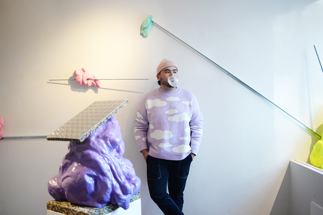 En proträttbild på konstnären Alejandro Montero Bravos som står lutad mot en vit vägg. Alejandro är iklädd lila tröja med moln på, en rosa mössa och svarta byxor. 