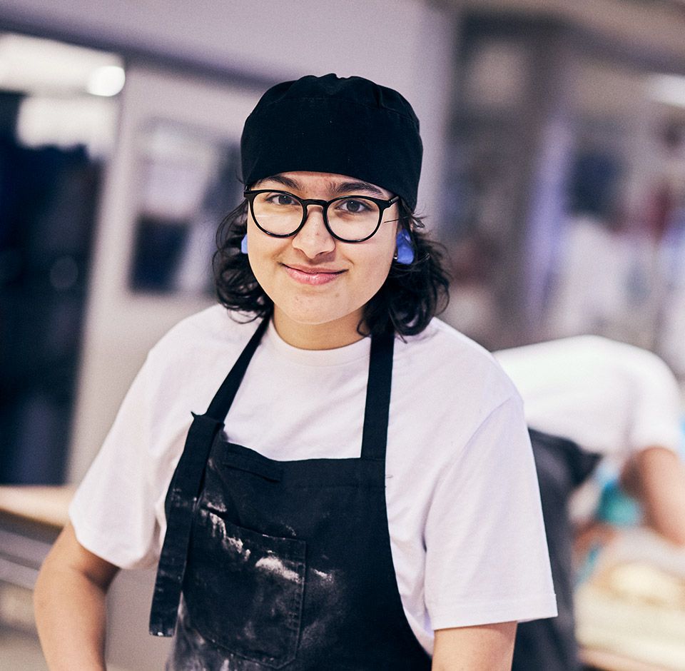En leende elev med kockmössa, glasögon och förkläde i bagerimiljö