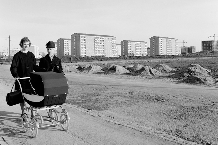 Svartvitt fotografi av två personer med barnvagn. I bakgrunden syns grushögar och några höghus