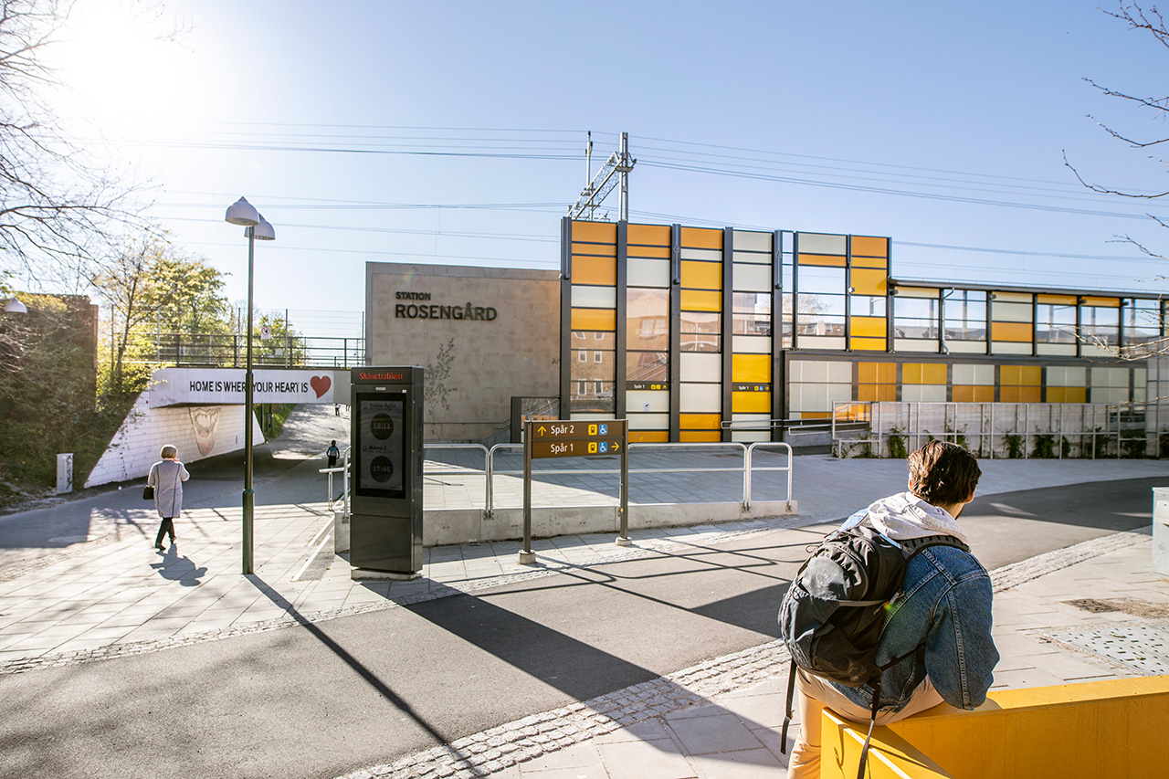 Station Rosengård en solig dag. TIll vänster en person på väg genom tunnel med texten "Home is where the heart is" upptill och till höger syns stationen i bakgrunden och en person i jeansjacka i förgrunden. 