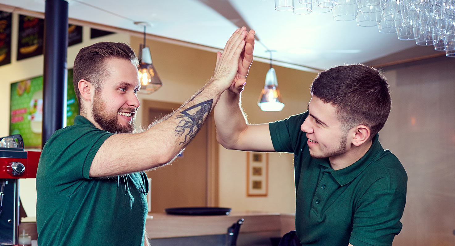 Två män i gröna tröjor som jobbar i en bar ger varandra en high-five.