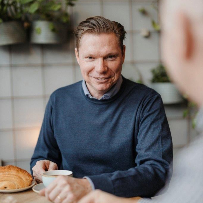 Jesper iförd blå tröja sitter vid ett bord med en kaffekopp i handen.