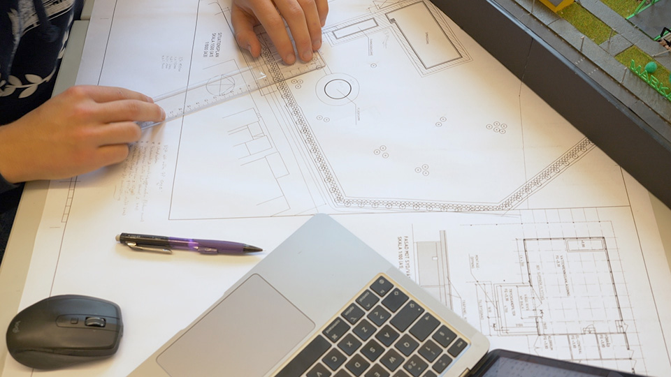 Närbild på skiss över en byggnad. På bilden syns också linjal, dator och två händer.