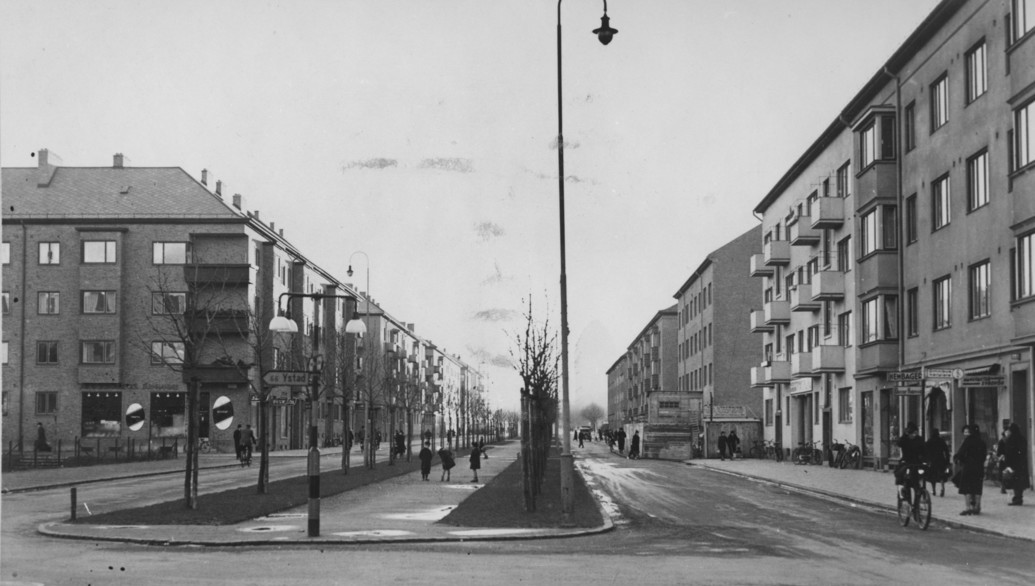 En historisk svartvit bild som visar en gata med människor och cyklar i rörelse.