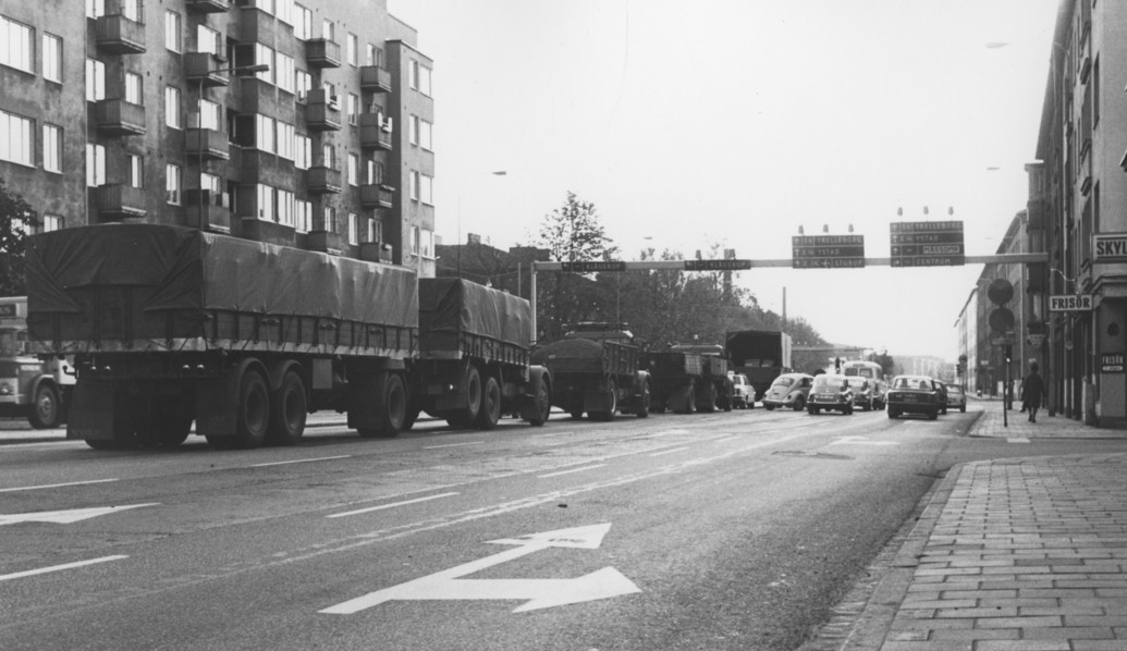 Svartvit bild på flerfilig väg med stora lastbilar och bilar som väntar vid trafikljus.