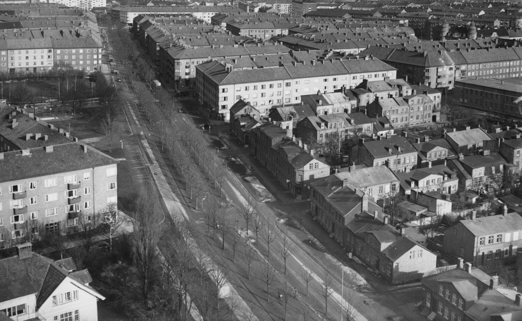 En gammal svartvit bild av en stadsgata med historiska byggnader och människor som promenerar.