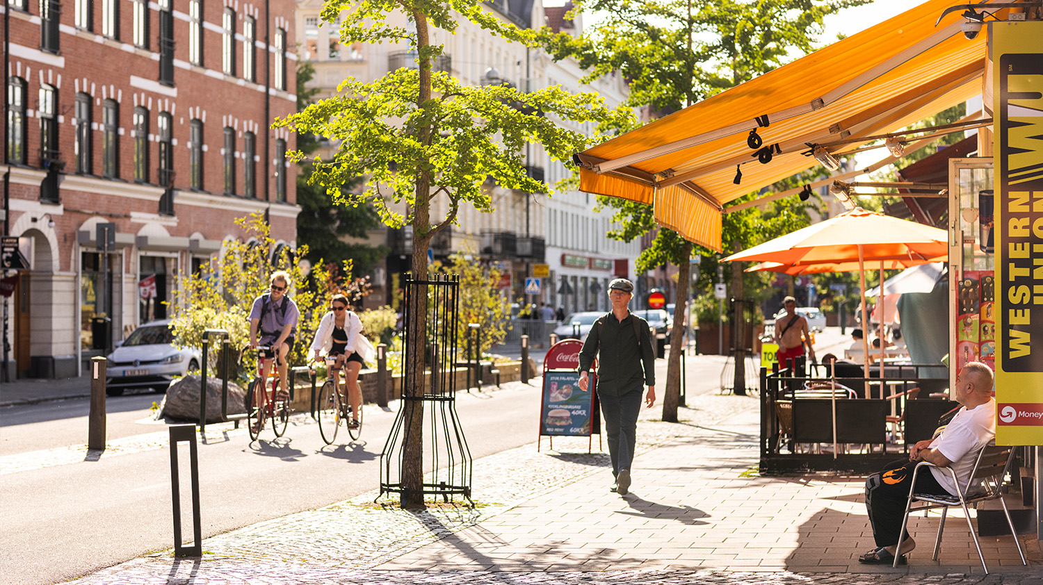 Fullt med liv på Ystadsgatan. En man sitter på en stol utanför en butik och tittar ut över förbipasserande på cykel och till fots.