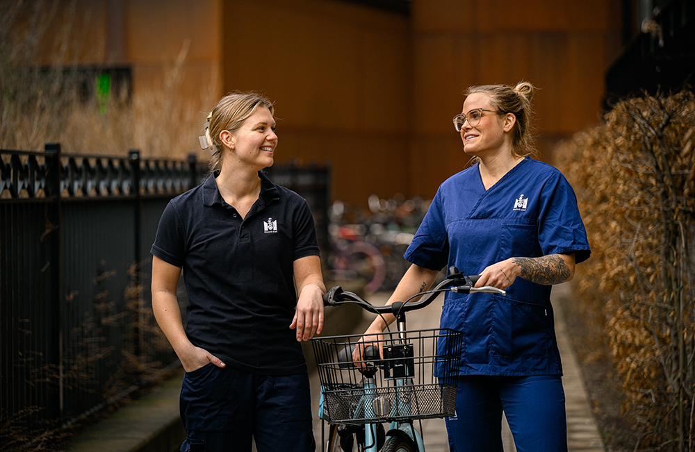 Två kvinnor i arbetskläder med Malmö stads logotyp står vid en cykel och tittar leende på varandra