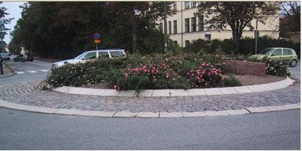 En rondell med körande bil. i mitten är det en yta för buskar, nedanför är det anlagd sten innan det blir en rondell med asfalt. 