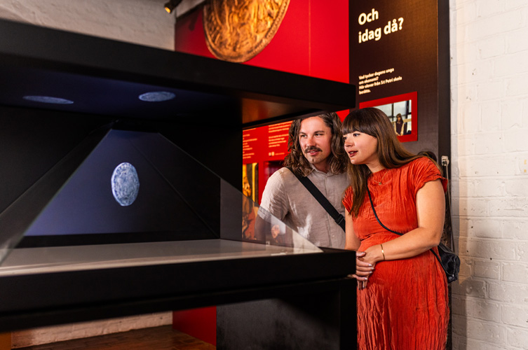 BIldspel med foto från utställningslokalen. MItt i står en stor box med ett hologram-mynt som snurrar runt. Det finns också en monter med föremål som man kan titta på genom ett förstoringsglas samt stora väggtexter med tillhörande bilder.