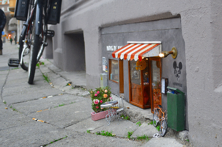 Litet minihus inbyggt i fasaden på bostadshus i centrala Malmö. Föreställer en litet restaurang med randig markis. 