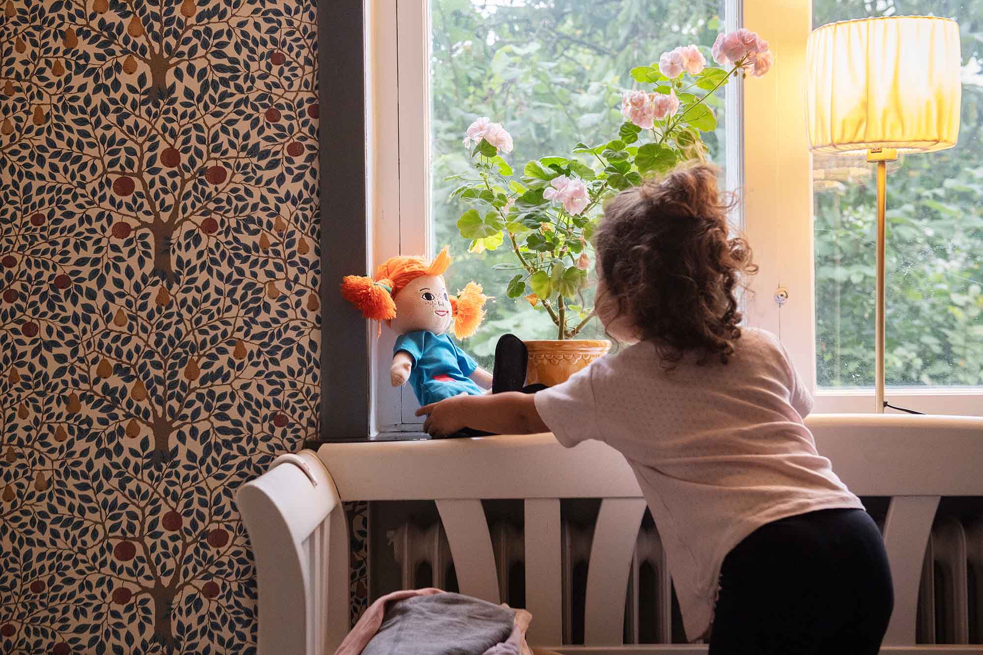 Barn leker med docka i hemmiljö, en fönsterlampa i förgrunden är tänd.