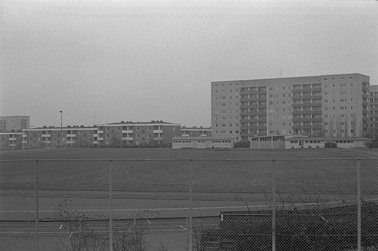Stor öppen idrottsplats, gräsbeklädd, i bakgrunden ett 9-våningshus och flera lägre 4-våningshus.