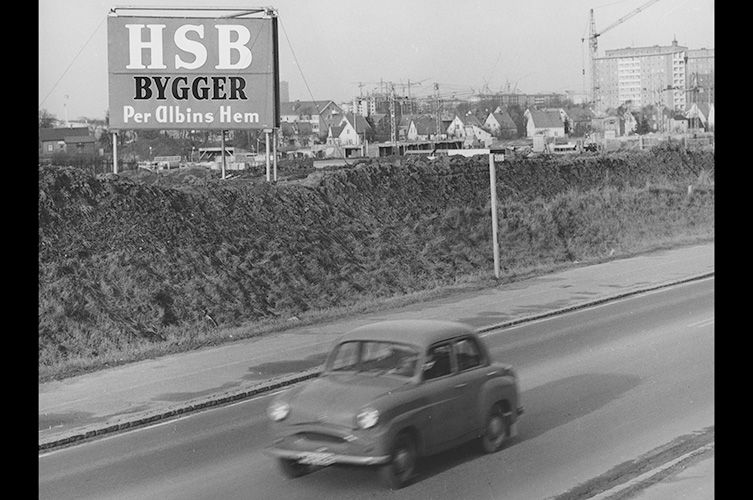 Äldre svartvitt foto med äldre Volvo från 60-talet i förgrunden och bostadsområde i bakgrunden.