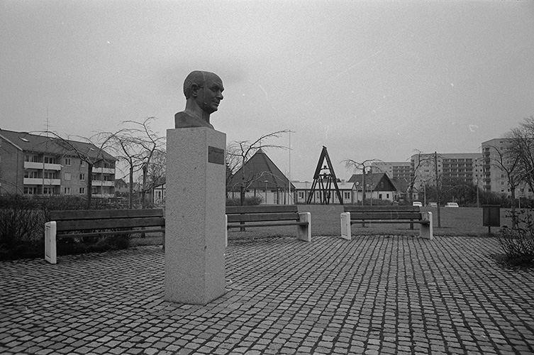 På torget står en byst upphöjd på en pelare, i bakgrunden syns bostadsområden med flerfamiljshus.