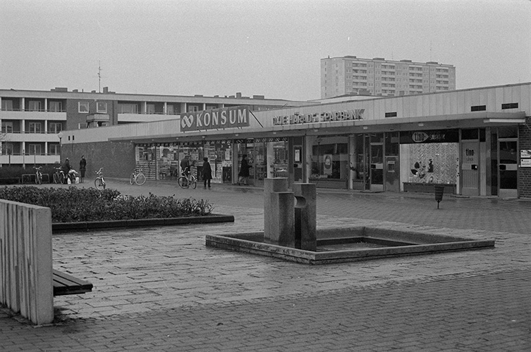 Stadsdelstorg med matvarubutiken Konsum och en tidstypisk stenskulptur på torget.