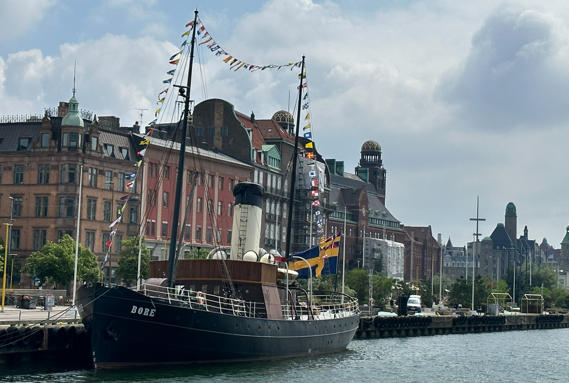 En svart båt med vit skorsten och färgglada flaggspel i två master ligger förtöjd vid en kaj.