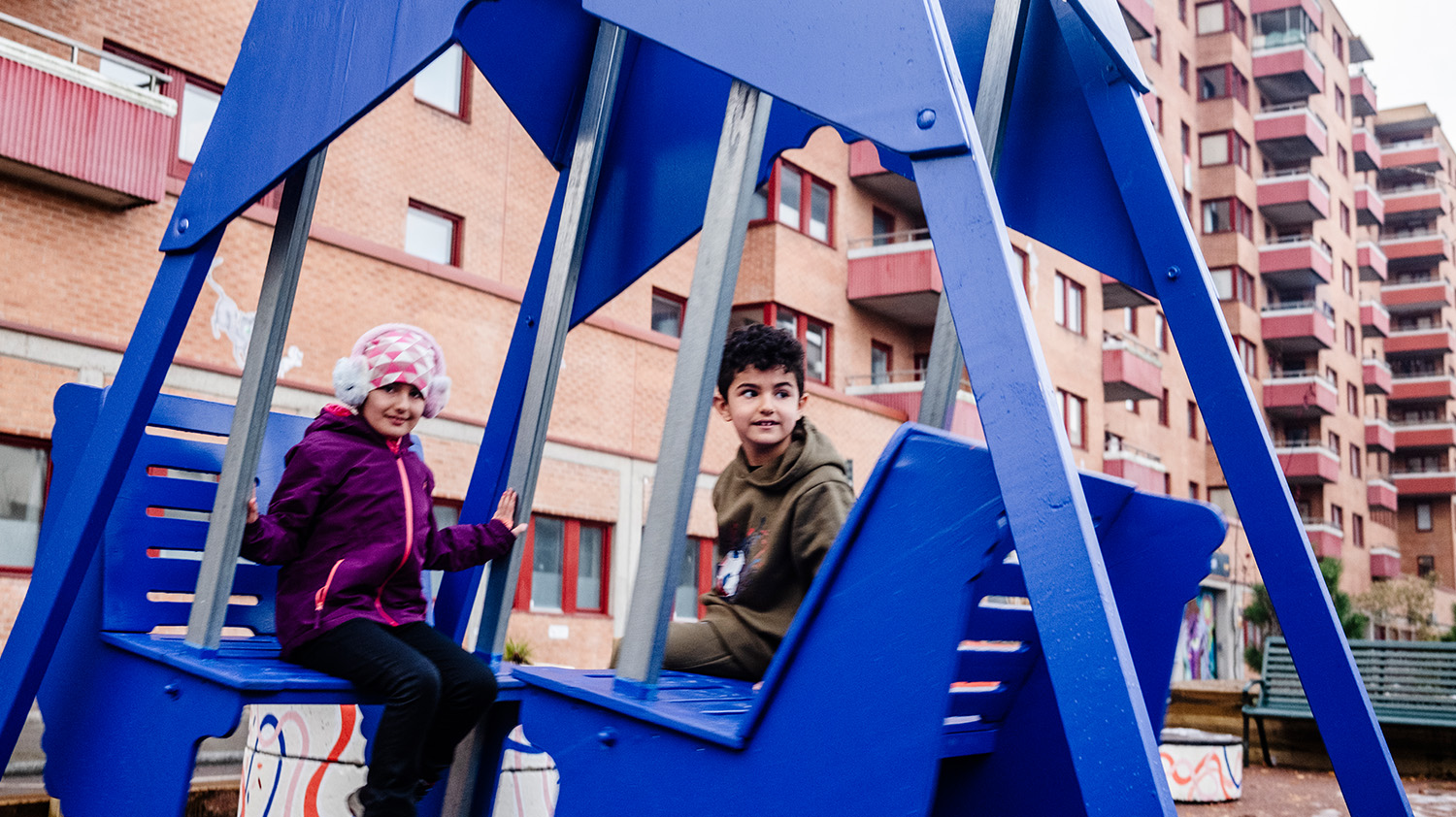 Två barn leker och ser glada ut i en blå gunga på den nya skolgatan på Friisgatan vintertid. 