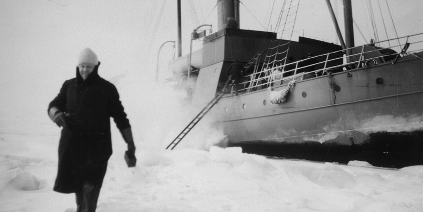 En historisk svartvit bild på en man som går på is och en båt i bakgrunden som stannat i isen.