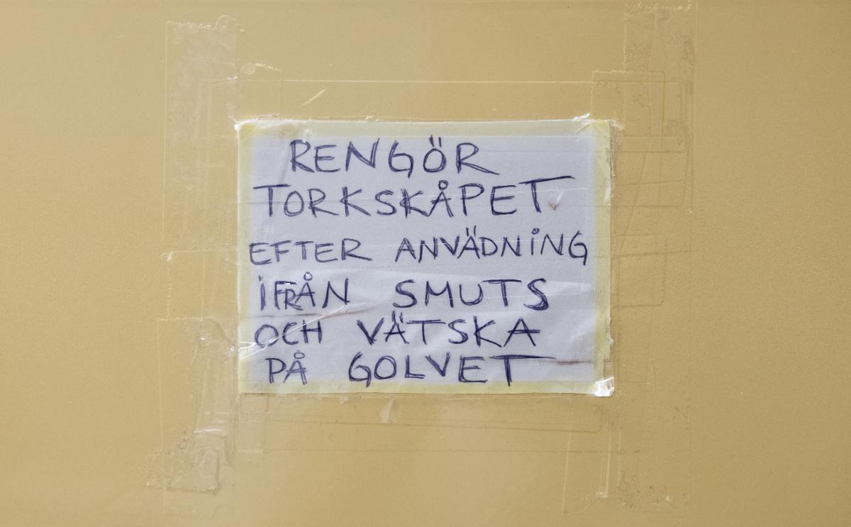 Vit papperslapp tejpad och limmad på torkskåp i gemensam tvättstuga. Lappen har texten ”RENGÖR TORKSKÅPET EFTER ANVÄDNiNG iFRÅN SMUTS OCH VÄTSKA PÅ GOLVET” skriven för hand med blå kulspetspenna.
