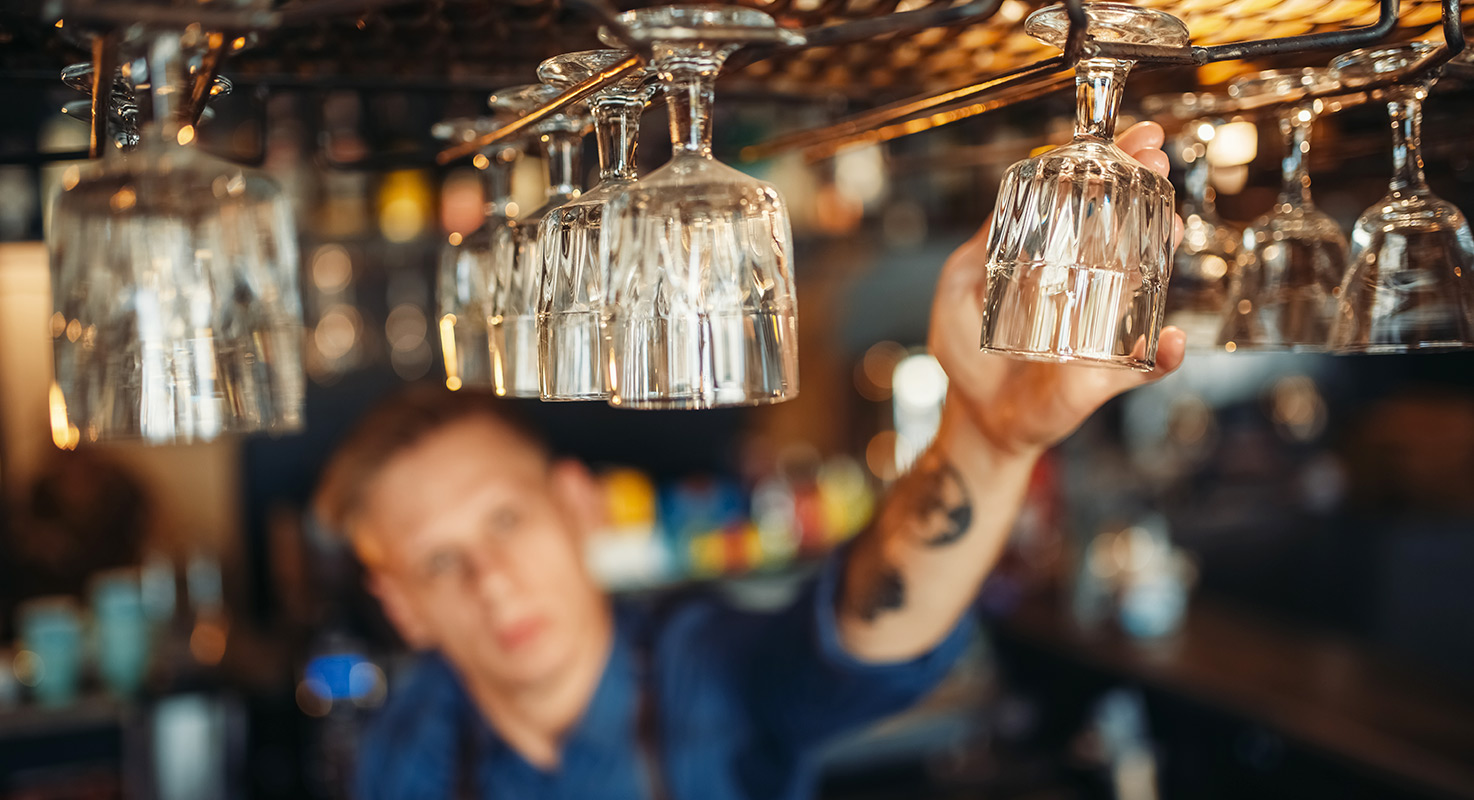 Anställd i baren sträcker sig efter vinglas som hänger i taket ovanför bardisken.