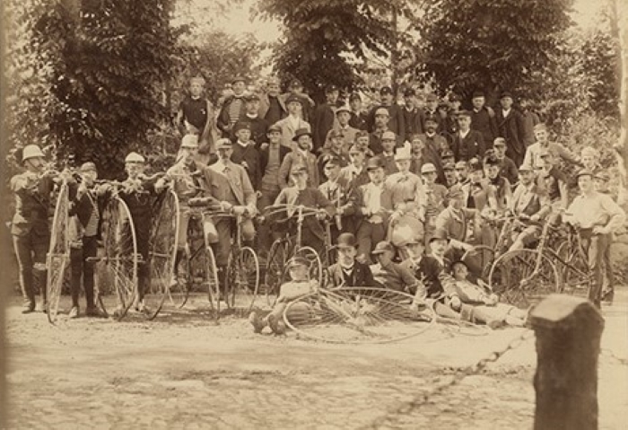 Gammalt lite gulnat fotografi av en grupp män som står uppställda med sina cyklar med stora framhjul.
