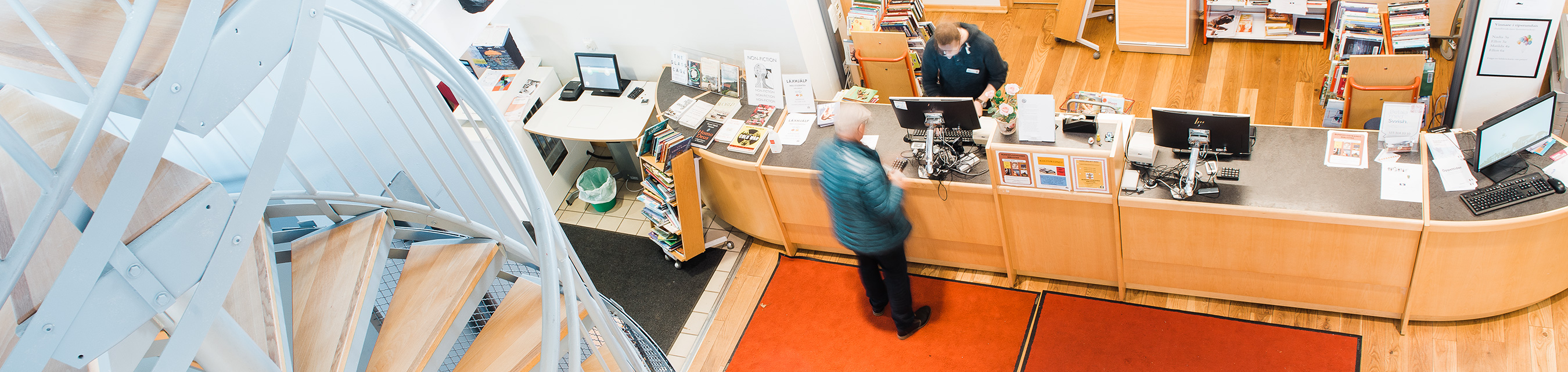 Interiör ovanifrån av Bunkeflostrands bibliotek. Personal hjälper en besökare vid informationsdisken.