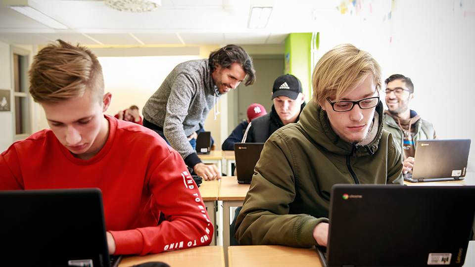 Flera elever arbetar med sina datorer i ett klassrum. En lärare instruerar en elev.