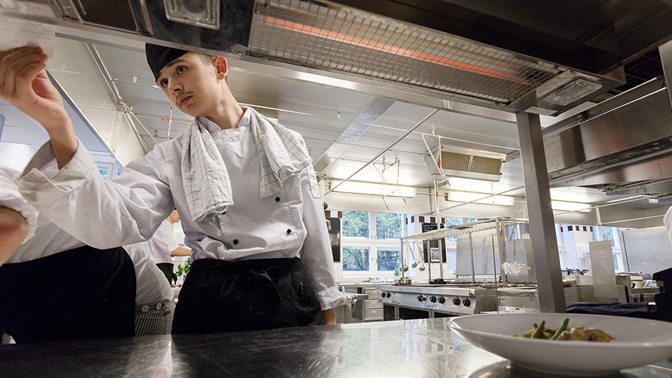 Ung man i kockkläder syns genom en öppen hylla i ett restaurangkök