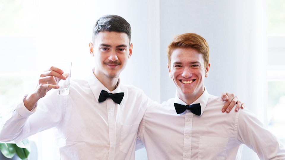 Två unga män i vit skjorta och svart fluga håller varandra runt axlarna och ler mot kameran . Mannen till vänste håller upp ett glas.