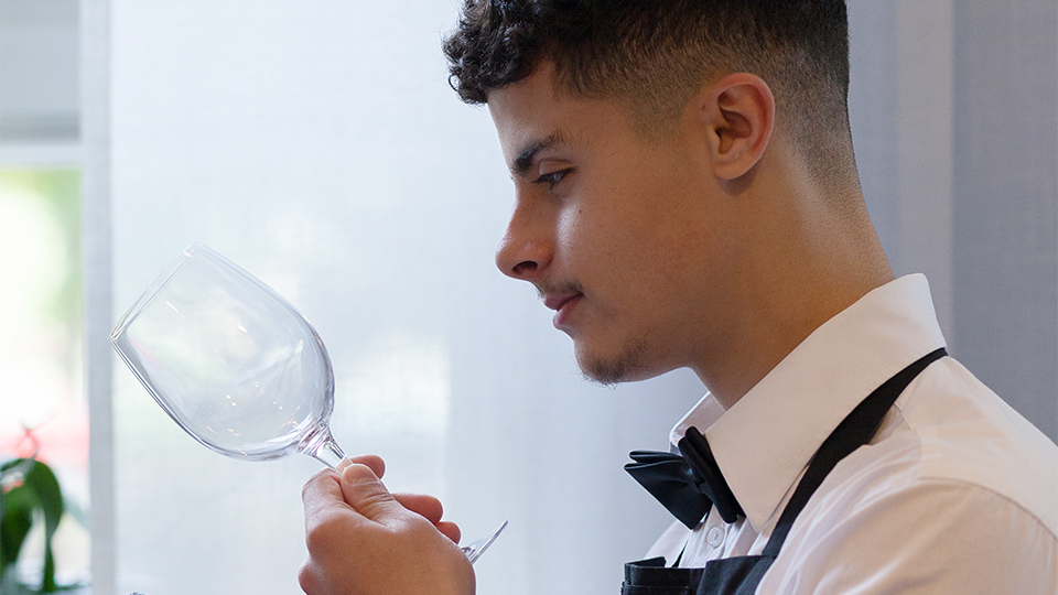 En servitörelev i vit skjorta, fluga och
förkläde studerar ett vinglas