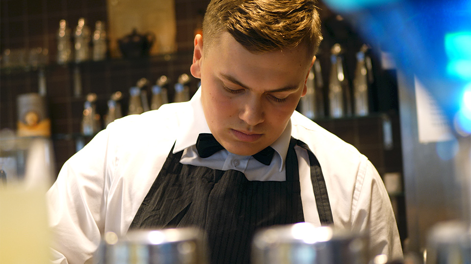 En ung man står bakom en bardisk och tittar ned mot sina händer.