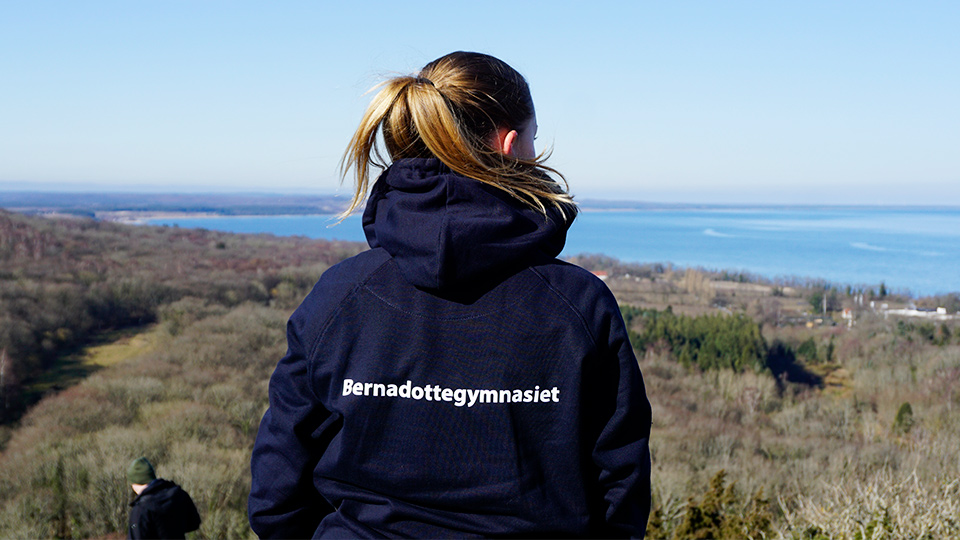 En elev står högt upp på ett berg och blickar ut över hav och natur. Vi ser ryggtavlan på eleven och på ryggen står texten Bernadottegymnasiet.