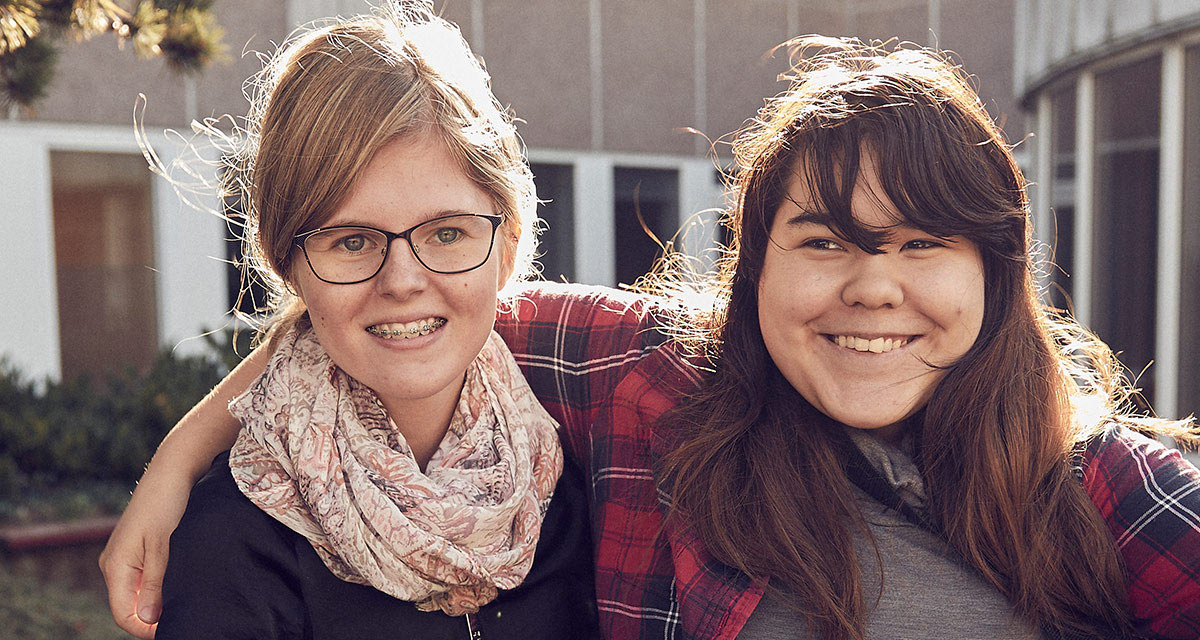 Två tjejer framför skolan. Den ena håller armen om axlarna på den andra.