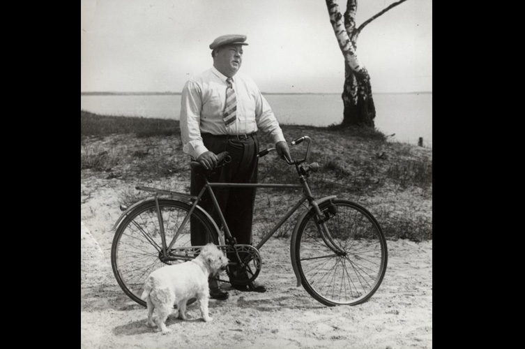 Äldre fotografi på Edvard Persson som står med en cykel på en strand. Framför cykeln intill Persson står en liten hund.