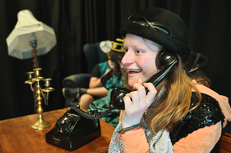 Ung leende kvinna sitter i en teatersal med gammaldags telefon mot örat. Bär teaterkläder.