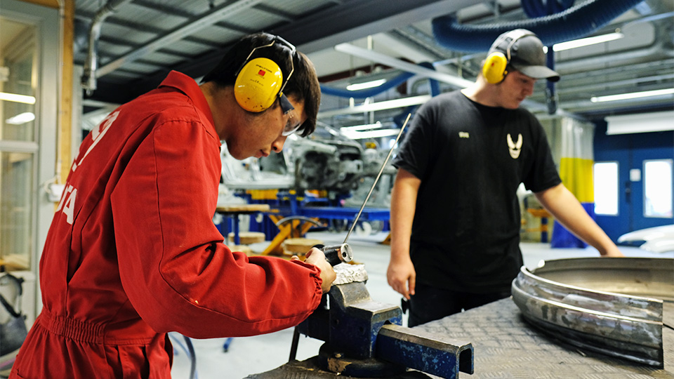 Två elever med hörselskydd slipar på bildelar.