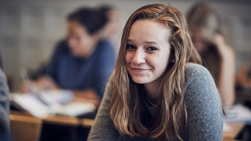 Glad tjej med långt hår och grå tröja i
klassrummet, ler mot kameran.