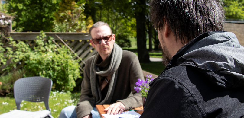 Två män samtalar vid ett bord utomhus.