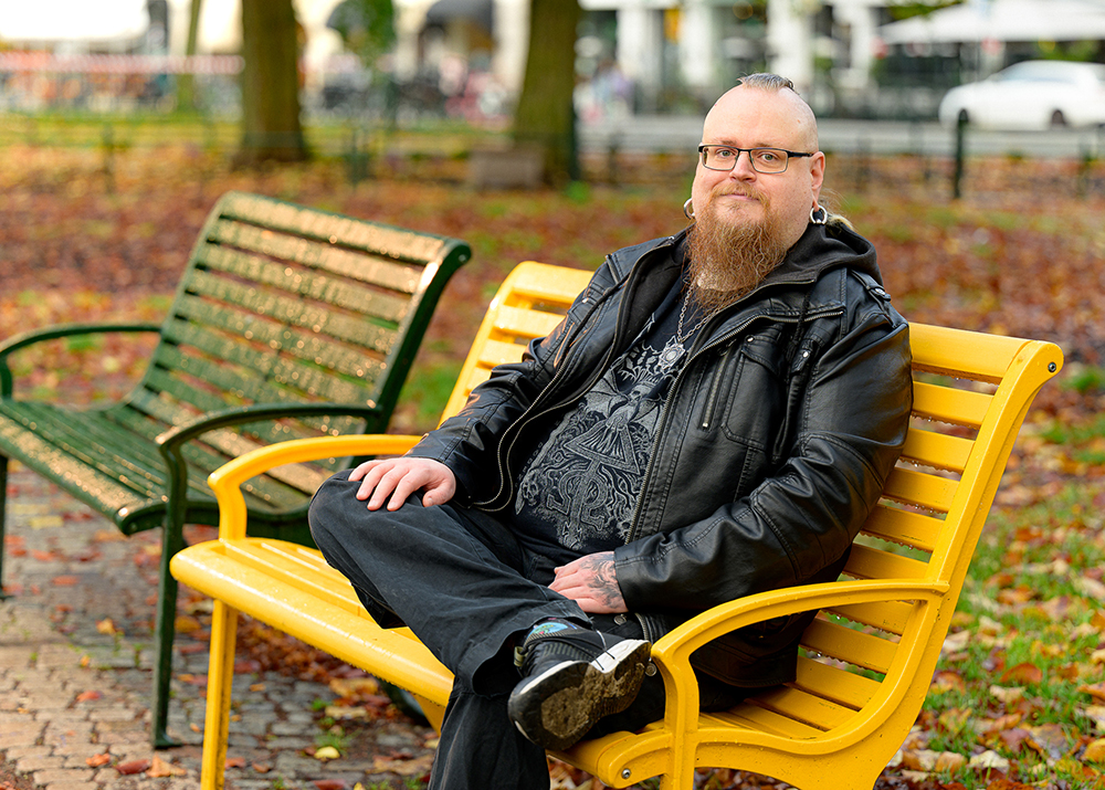 Man med glasögon, skägg och svarta kläder sitter på en gul vänskapsbänk och tittar in i kameran.