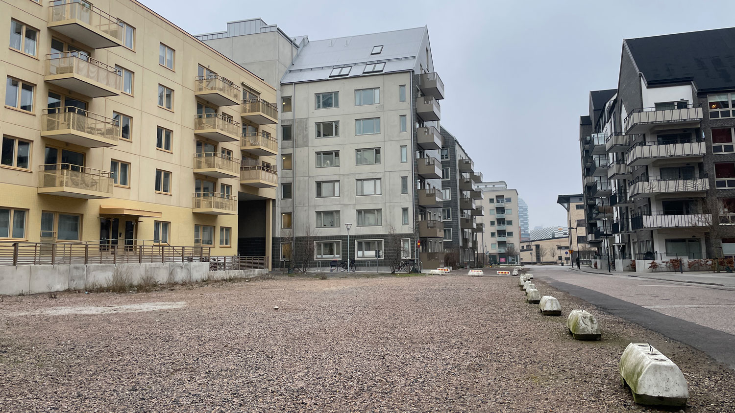 En grusad yta intill moderna bostadshus med guldgula och mörkgrå fasader, och låga betongklumpar som spärrar av mot gatan intill.