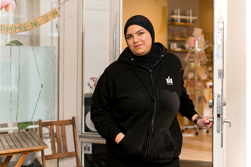 Kvinna med svart sjal och svart hoodie med Malmö stad-logga småler in i kameran och håller upp en dörr