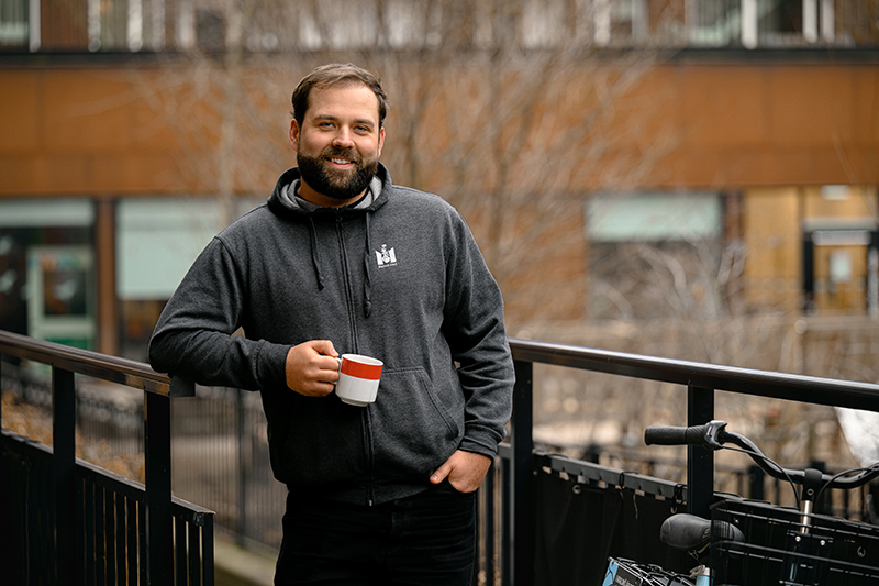 Man med brunt hår och skägg och grå hoodie med Malmö stad-logga håller en kaffekopp i handen och ler in i kameran