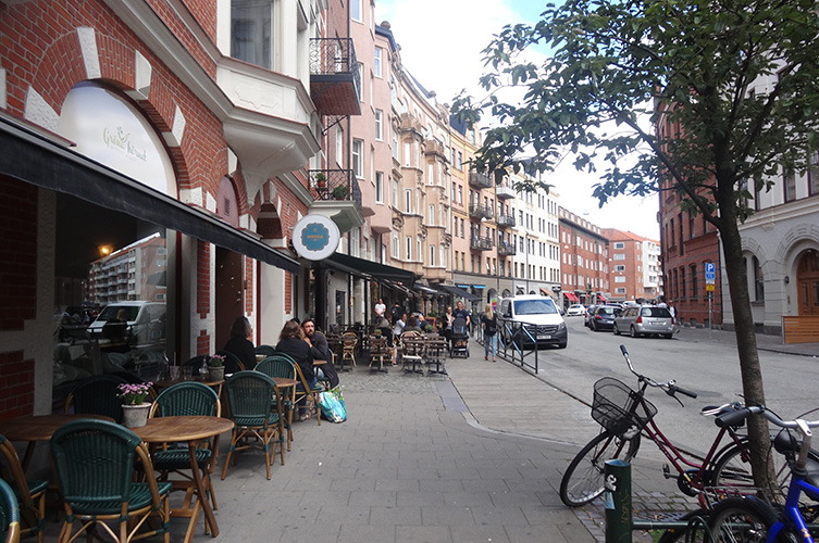 Del av gata med trottoar, träd, kafé med markiser utfällda,
och cyklar parkerade.