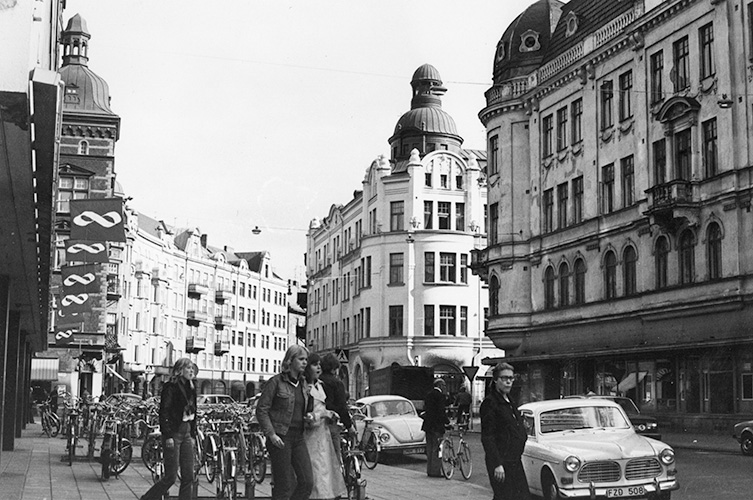 Svartvitt foto av gata med ståtliga äldre byggnader, folkliv samt parkerade cyklar och bilar.
